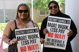Two black women in sunglasses, one with a headscarf, hold signs saying ‘More work, less pay, NO WAY’
