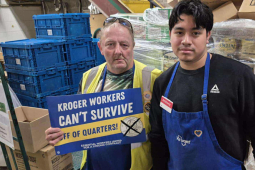 Two Kroger workers stand in front of pallets. One holds a sign that says "Kroger workers can't survive off of quarters."
