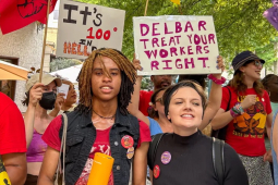 Several people with colorful signs in red and orange face the camera, one says “Delbar treat your workers right”