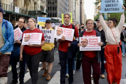 Eight people walk towards the camera, three with signs that say, “shame, Russel Vought”