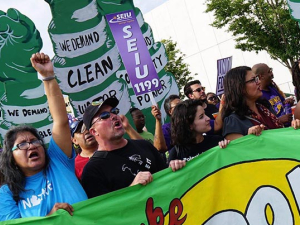 Crowd at rally behind bright green banner, woman with fist up, people holding signs, one SEIU sign visible, another for racial justice, picture at an angle