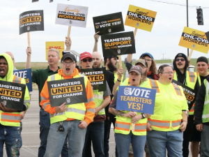 A group of 15 or so workers in hi-viz vests look excited and hold signs about voting for the Teamsters union.