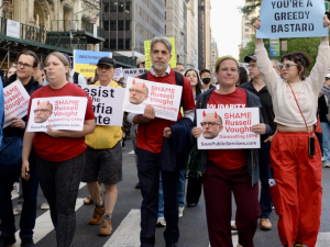 Eight people walk towards the camera, three with signs that say, “shame, Russel Vought”