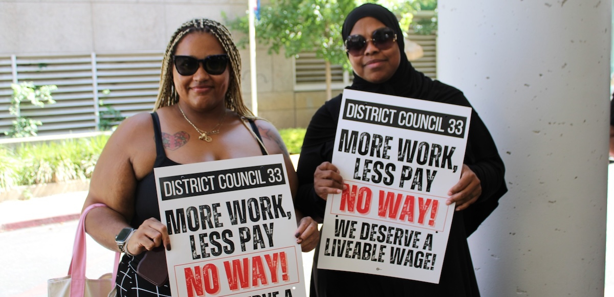 Two black women in sunglasses, one with a headscarf, hold signs saying ‘More work, less pay, NO WAY’