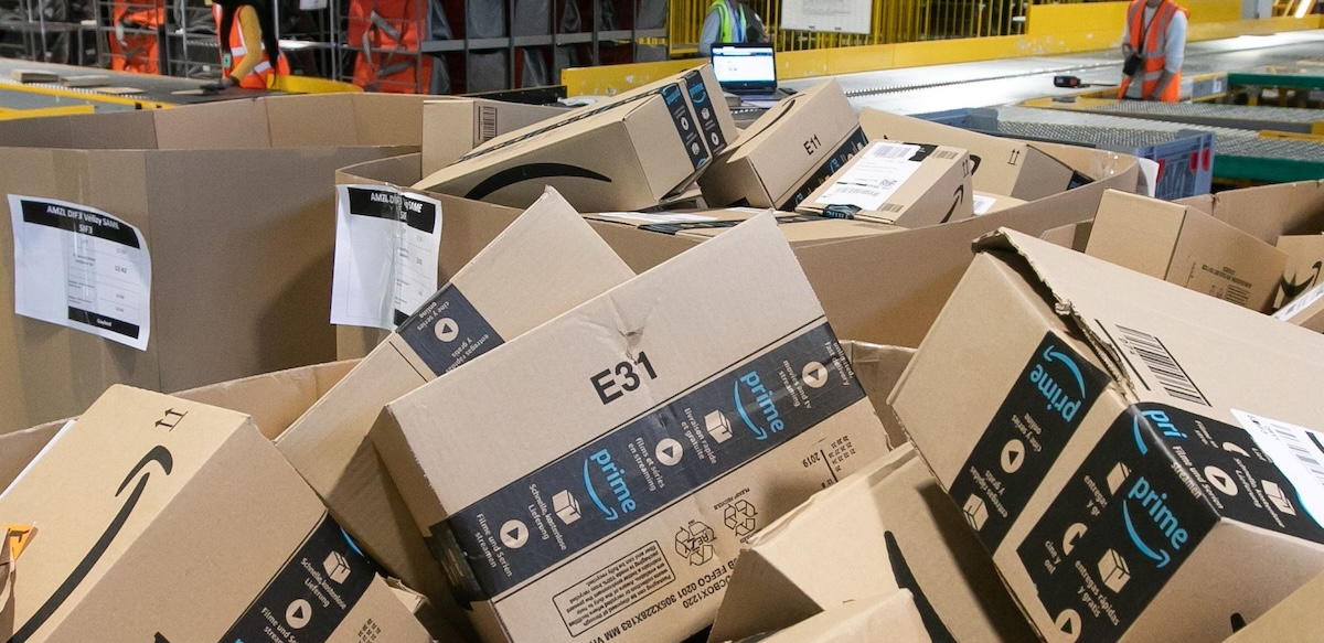 A pile of Amazon boxes is shown with workers small in the background