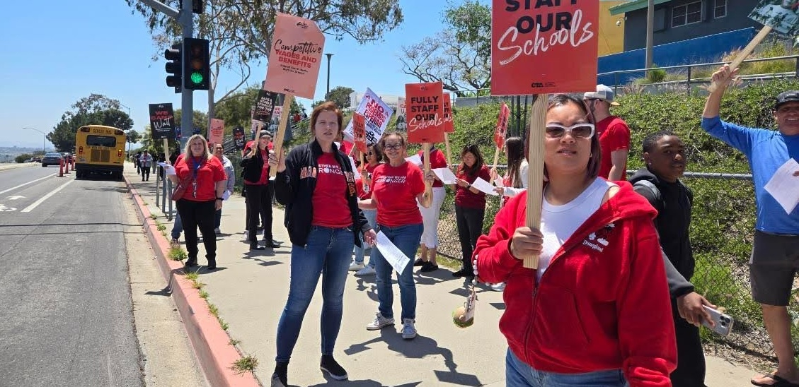 Teachers in red picket alongside a road with “Fully Staff Our Schools signs”