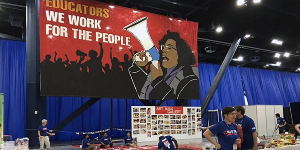 A banner says “Educators, we work for the people” in the background as union members are visible setting up for a meeting in the foreground.
