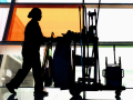 A woman custodian pushes a cart full of implements in profile past colorful windows.