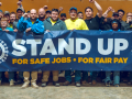 Thirty people, many with fists raised stand behind a banner that says “Stand Up, for safe jobs, for fair pay”