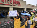 Workers in yellow shirts and rain parkas picket in front of a grocery store with a ‘stop the layoffs’ signs visible