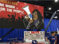 A banner says “Educators, we work for the people” in the background as union members are visible setting up for a meeting in the foreground. 
