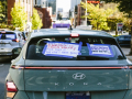 A car back window carries signs, including one saying, “Would you drive 10 miles for $4? Raise the Pay!”